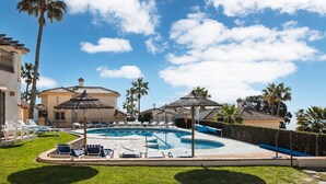 Una piscina con lettini e ombrelloni, circondata da palme e un edificio con tetto in tegole.