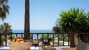 Un tavolo con una ciotola di frutta, bicchieri da vino e patatine, con vista panoramica sul mare e sulle palme.