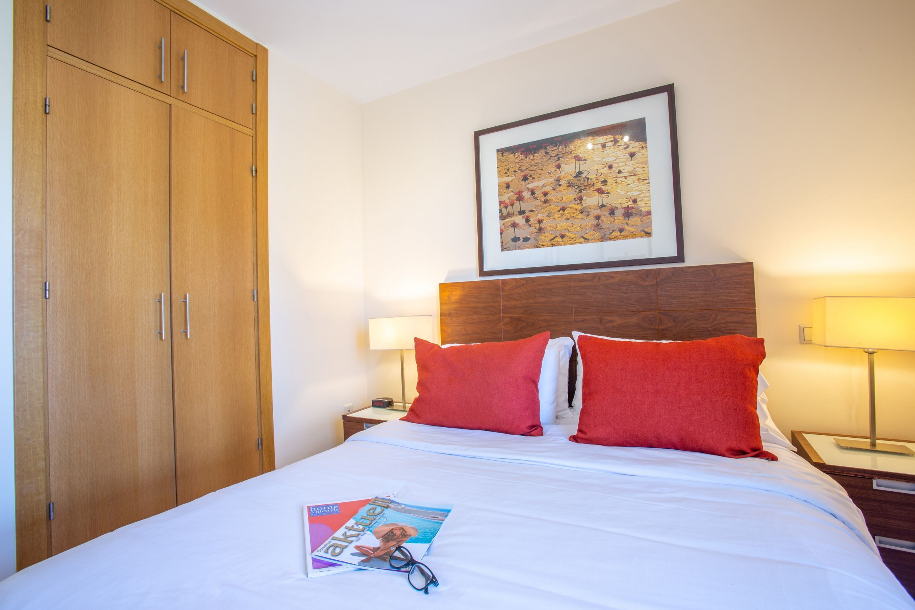 A hotel room with a bed, a wooden wardrobe, a framed artwork on the wall, and a magazine on the bed.