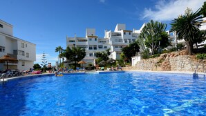 En swimmingpool med solstolar och parasoller, omgiven av byggnader med balkonger och palmer.