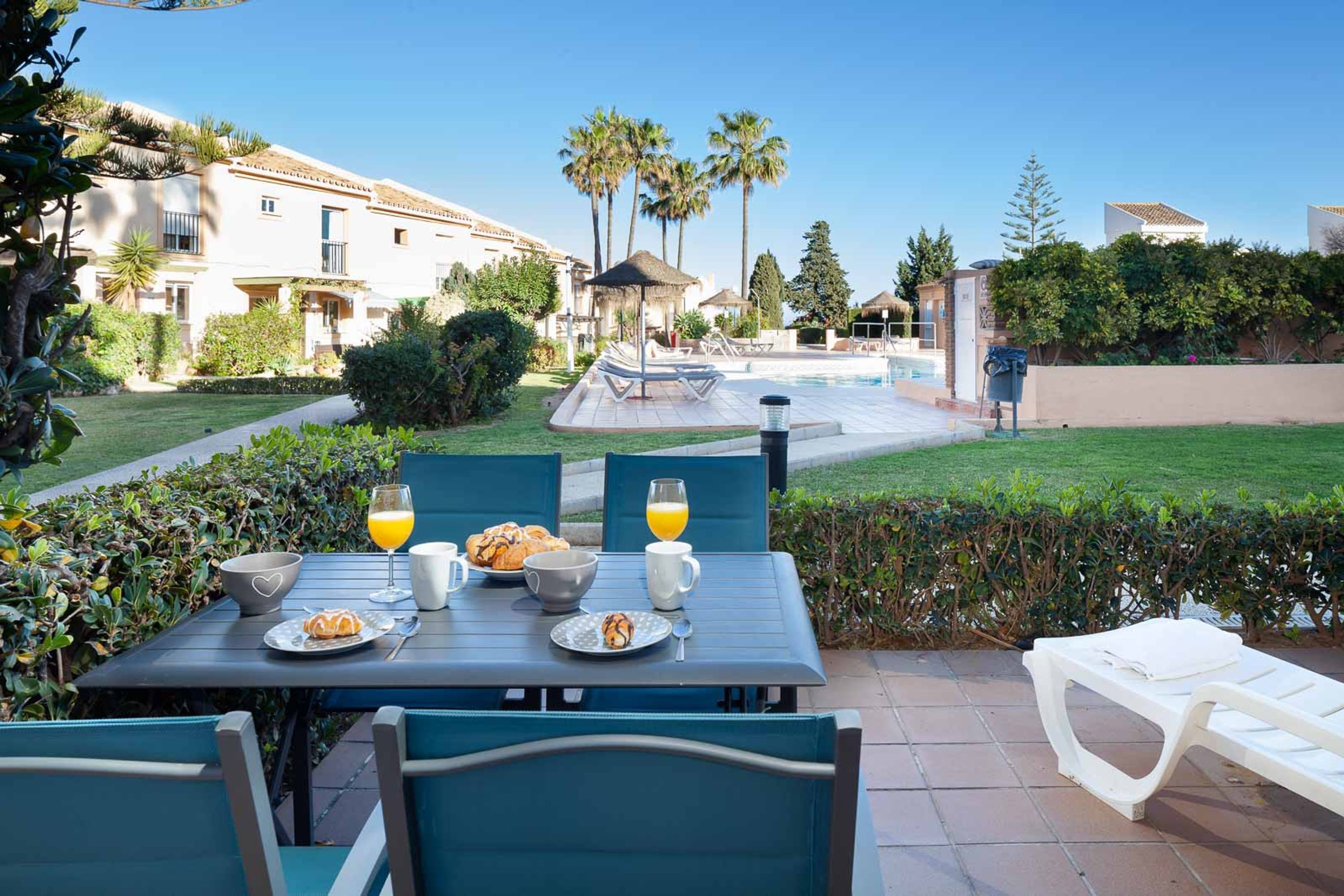 A patio with a table set for breakfast, a swimming pool, and a view of the surrounding buildings.