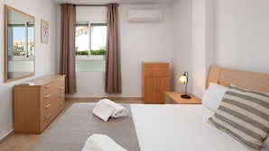 Una habitación de hotel con cama, cómoda de madera, mesita de noche con lámpara, armario y ventana con cortinas.
