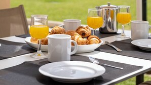 Ulkokäyttöön tarkoitettu ruokapöytä, jonka päällä on aamiaisherkkuja, kuten croissantteja ja appelsiinimehua, ja pöydän yläpuolella on suuri aurinkovarjo.