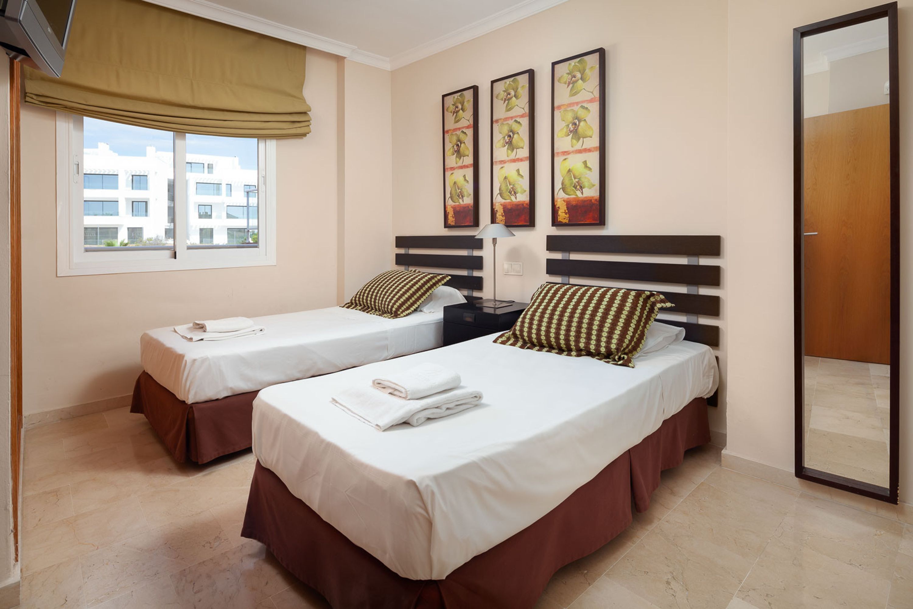 Une chambre d’hôtel avec deux lits, une fenêtre donnant sur des immeubles et trois tableaux encadrés accrochés au mur.