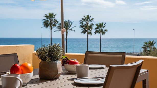 A table with a laptop, a bowl of fruit, a potted plant, and a white mug, overlooking the ocean with palm trees in the background. - AS MDM 5 in Mijas Costa (Mijas)