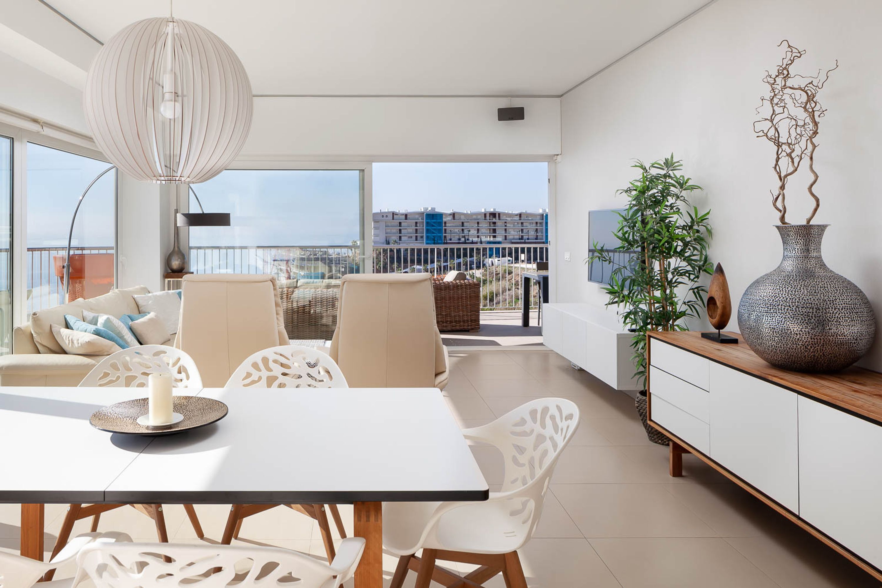 A modern living room with a dining table, chairs, a sofa, a large window, and a balcony with a view of buildings.