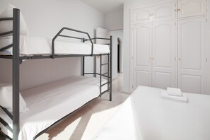 Een slaapkamer met een stapelbed, een wit bed, een klein tafeltje met een kussen en een kledingkast met deuren.