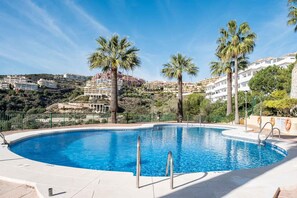 Een zwembad met helderblauw water, omringd door een hek, palmbomen en gebouwen op de achtergrond.