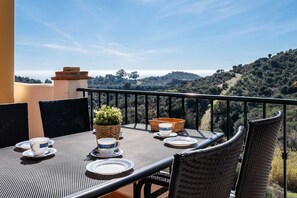 Een balkon met een tafel voor twee personen, stoelen, een bloempot en uitzicht op een glooiend landschap.