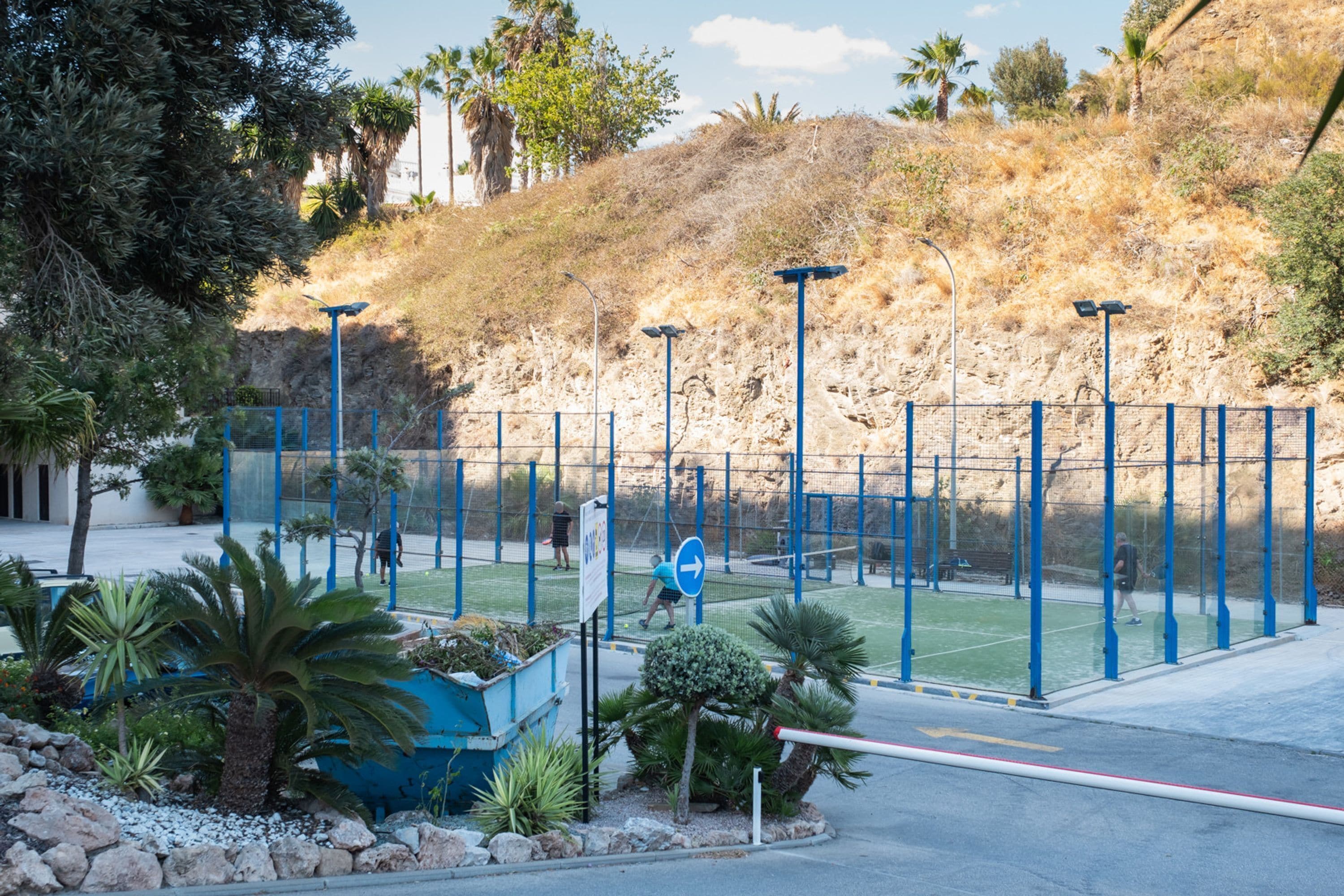 Een omheind tennisveld met een blauw net, omgeven door palmbomen en een rotsachtige helling.