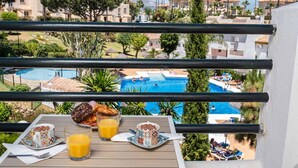 Balcón con vista a una piscina, mesa de comedor con productos para el desayuno y zona junto a la piscina con sillones de descanso.