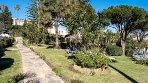 Un jardín con un sendero de piedra, árboles y una zona de piscina con sillones de descanso.