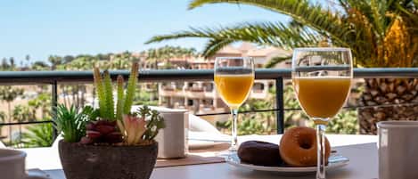 Una mesa con un vaso de jugo de naranja, un donut y una taza de café, con vista a palmeras y edificios.