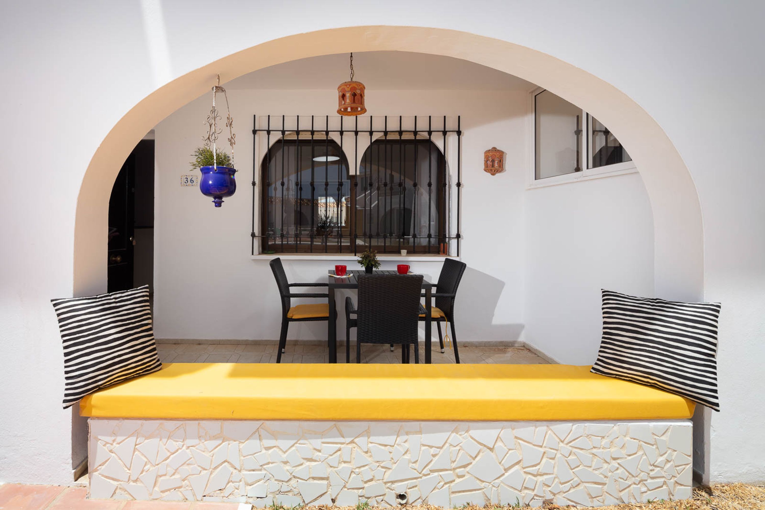 A room with a white wall, a yellow bench, a table with chairs, and a window with a metal grill.