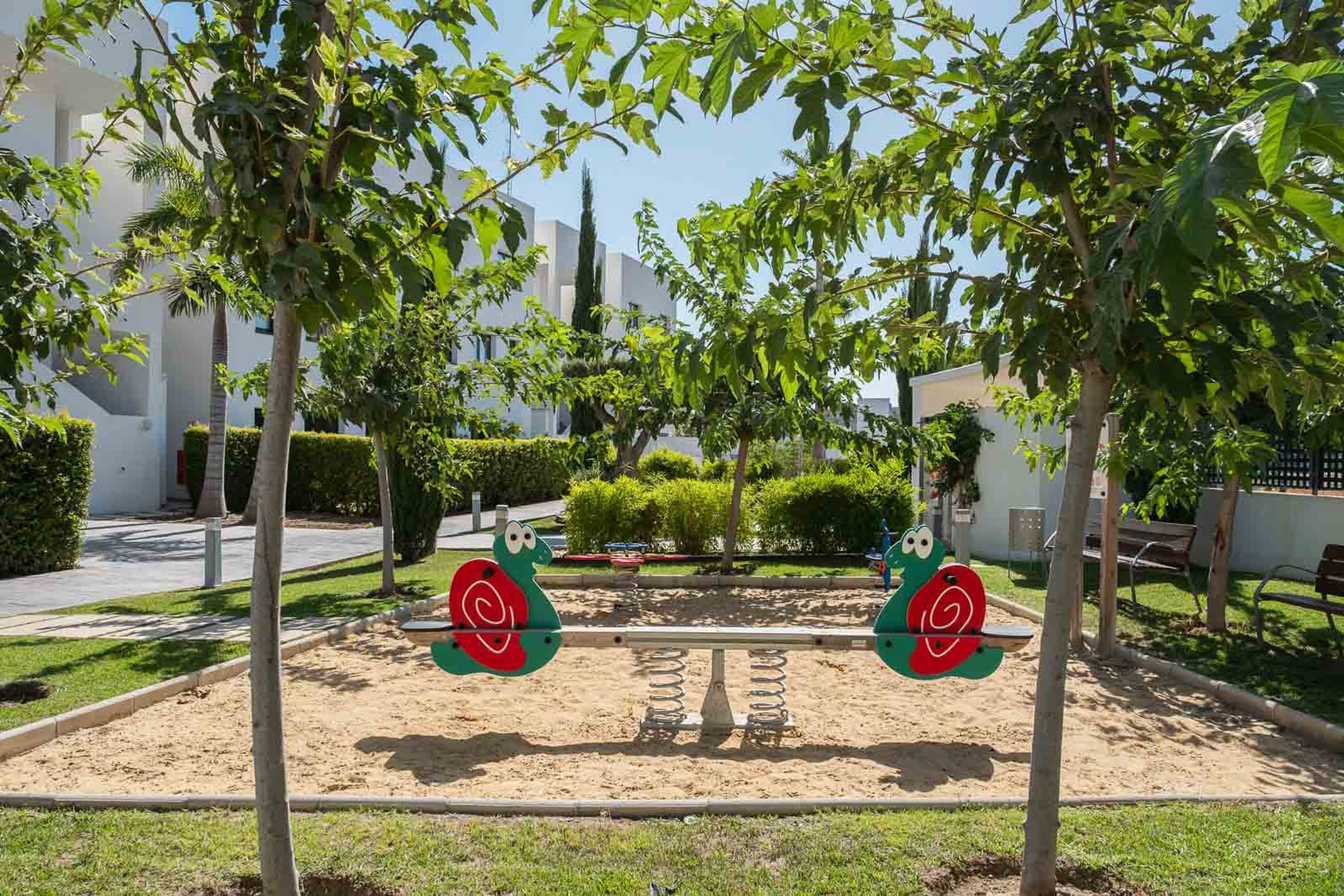A playground with a seesaw and swings, surrounded by trees and a building in the background.