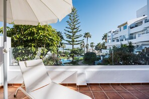 Un balcón con una silla blanca, un toldo grande y vistas a una piscina y edificios.
