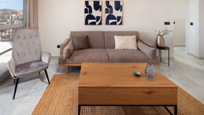 Uma sala de estar com sofá, uma poltrona, uma mesa de centro, arte abstrata na parede, um vaso com flores e vista pela janela.