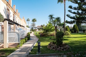 Un chemin menant à un bâtiment à la façade blanche, avec un jardin agrémenté de palmiers et d’arbustes.