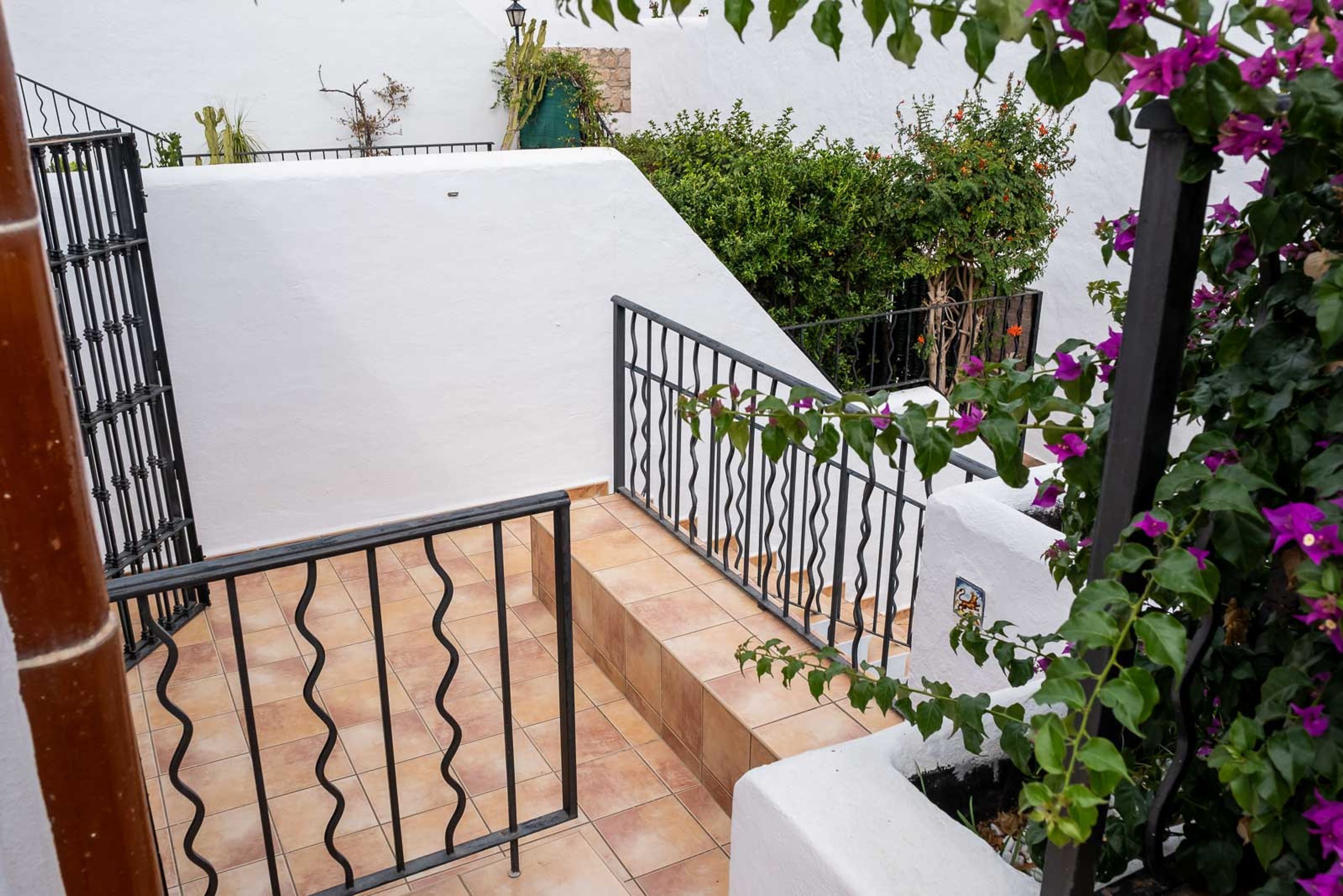 Un edificio blanco con escaleras, barandilla de metal y un jardín con plantas verdes y flores moradas.