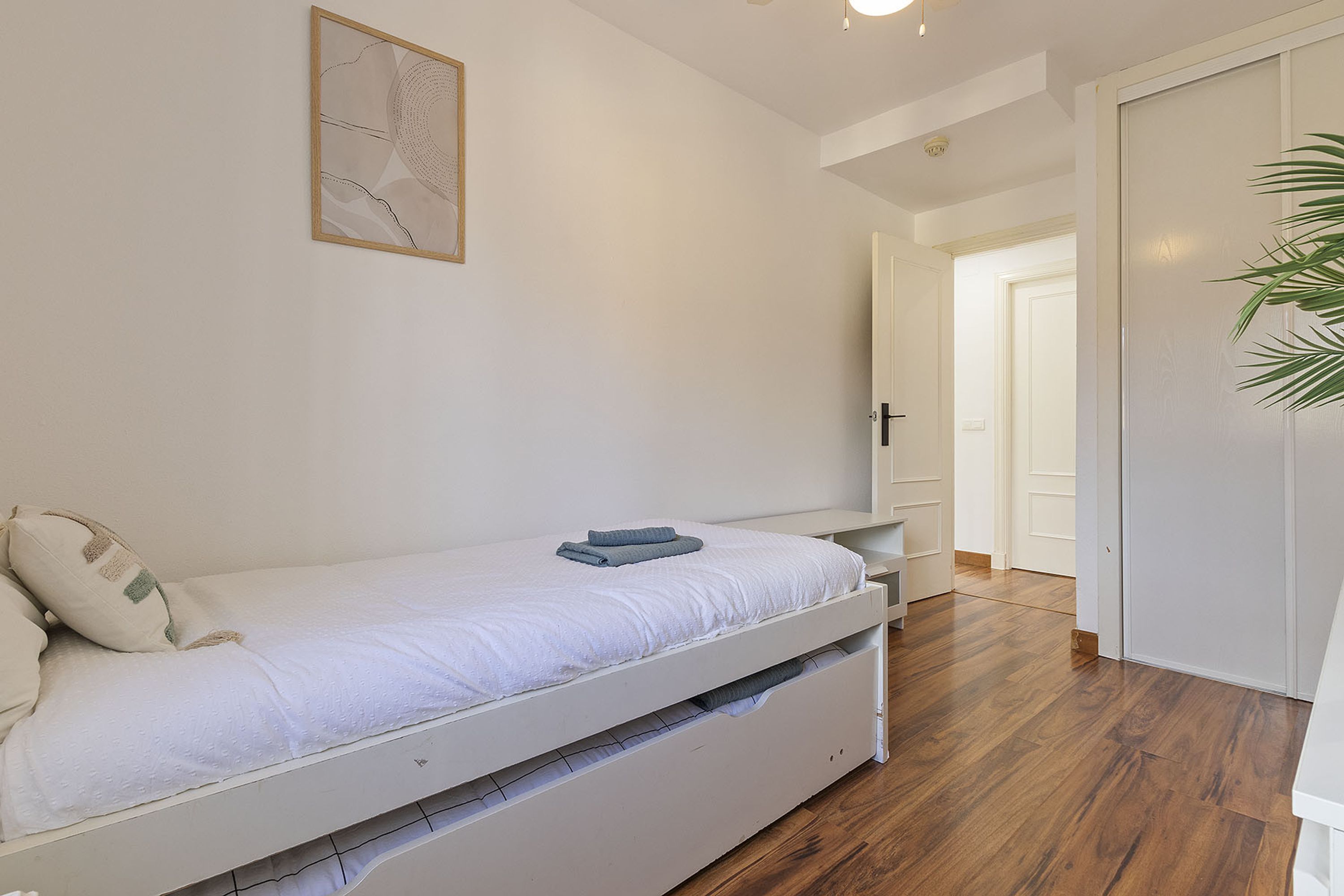 A single bed with a white mattress and a blue towel, a wooden floor, and a door leading to another room.