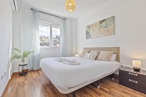 Un dormitorio con una cama grande, mesitas de noche, una planta en maceta y una ventana con cortinas.