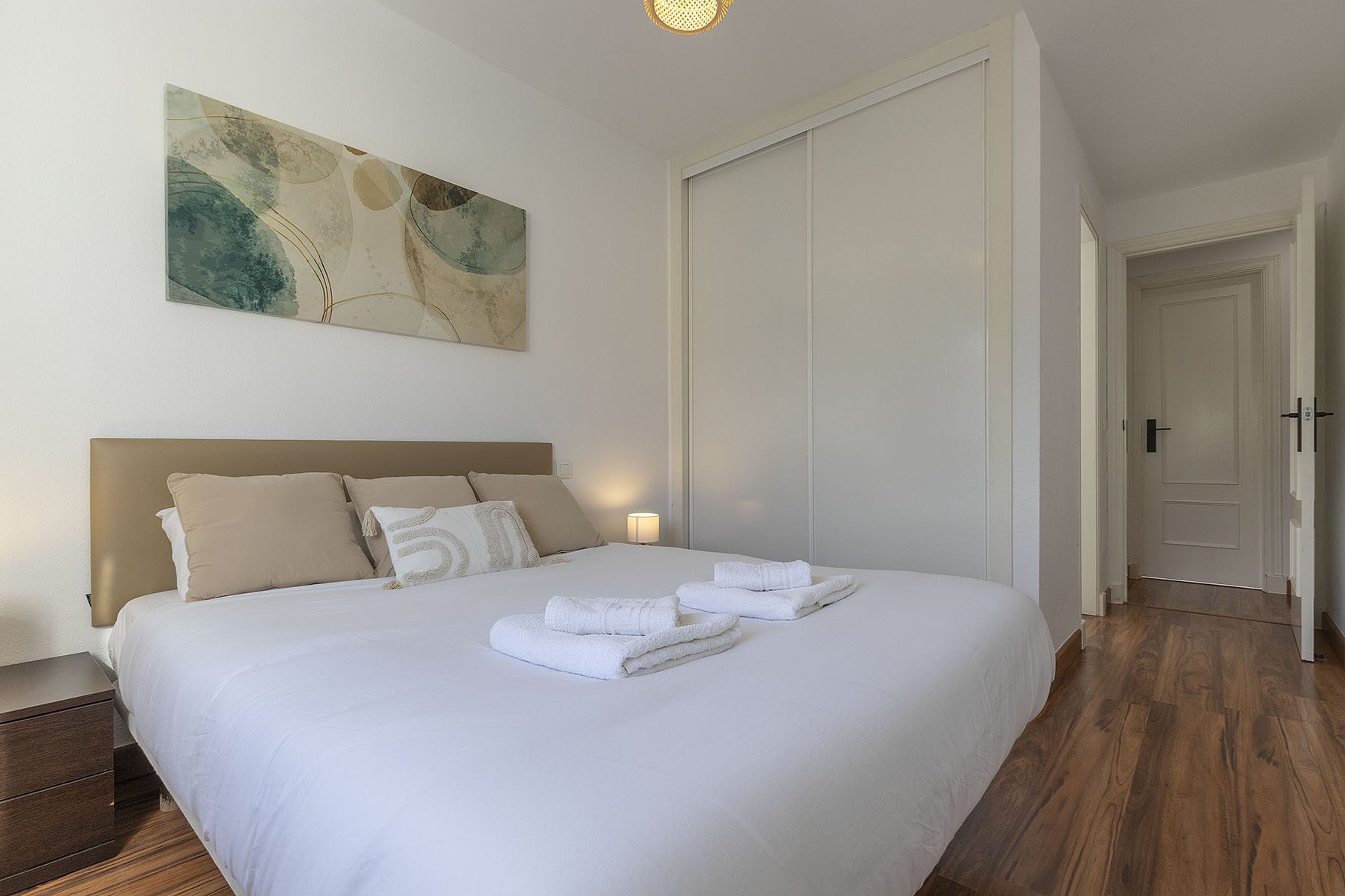 A neatly made bed with white linens and towels, a headboard, a wall-mounted abstract painting, and a wooden floor.