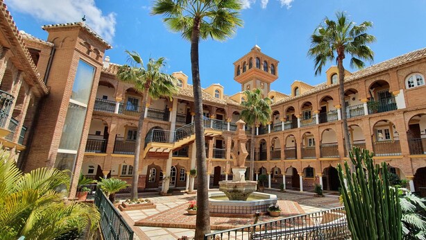 A courtyard with a fountain, palm trees, and a building with multiple balconies and arches. - AS EXT Mijas Puebla Aida 12L (Mijas)