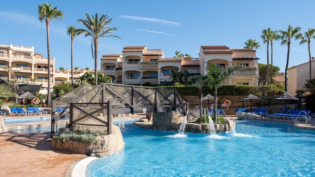 A swimming pool with a waterfall feature, surrounded by palm trees and a building with multiple balconies. - AS MDM 3 in Mijas Costa (Mijas)