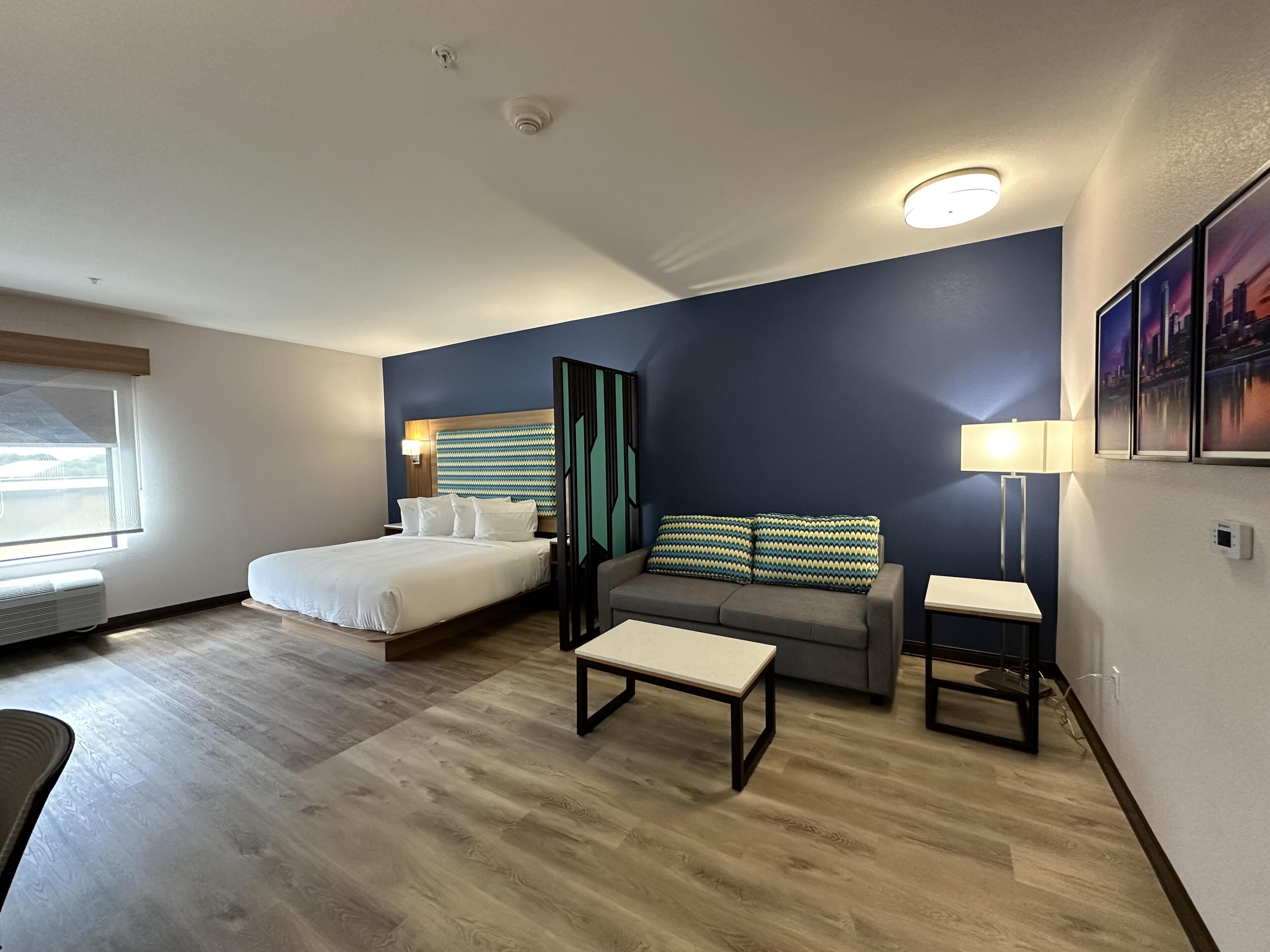 Ein Hotelzimmer mit einem Bett, einem Sofa, einem kleinen Tisch, einem Sessel, einem Fenster mit Jalousien und einer Wand-Klimaanlage.