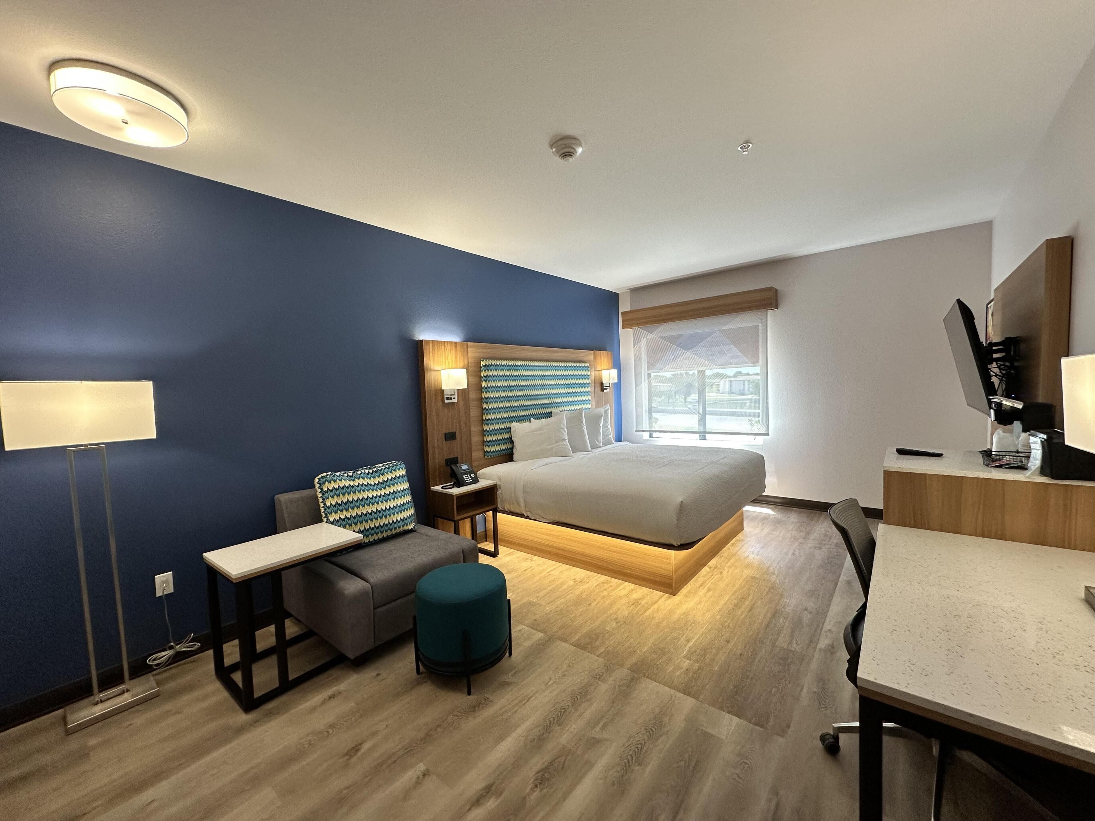 Una habitación de hotel moderna con cama, escritorio, silla, mesita de noche, lámpara y ventana con vistas.
