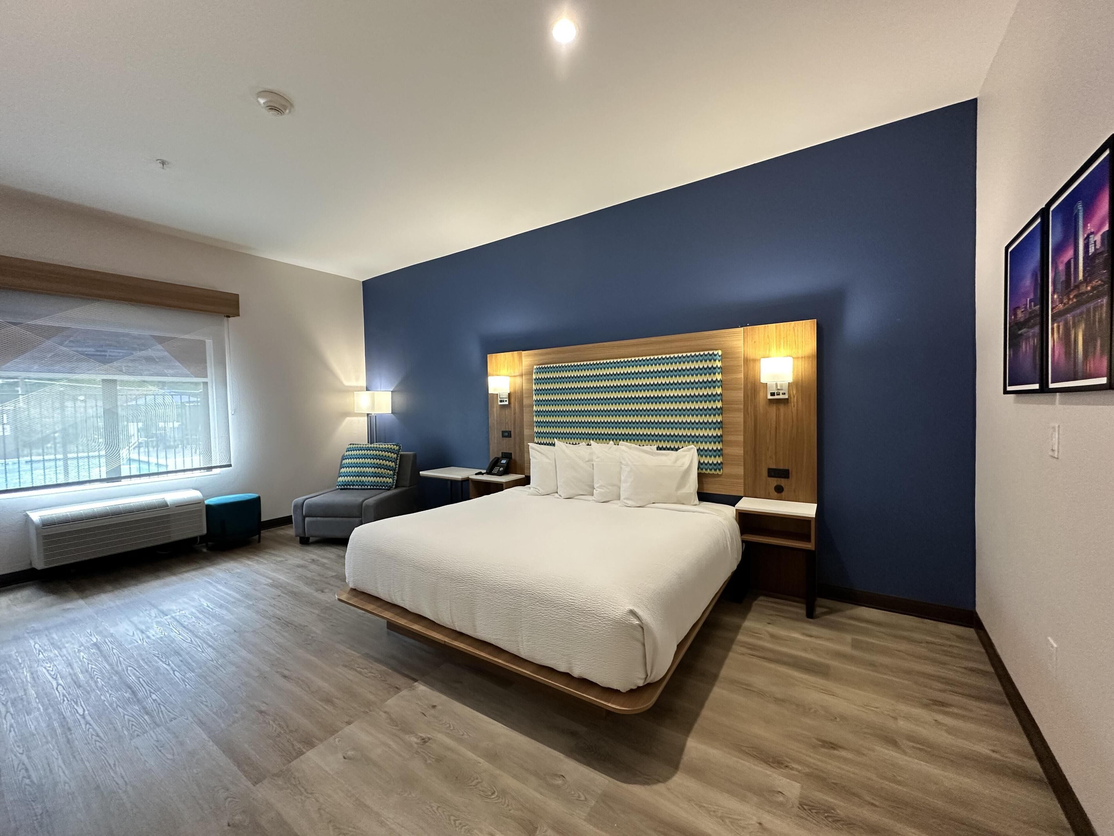 A modern hotel room with a large bed, a seating area with a chair and a small table, a wall-mounted TV, and a window with a view of a building.