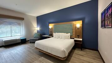 A modern hotel room with a large bed, a seating area with a chair and a small table, a wall-mounted TV, and a window with a view of a building.