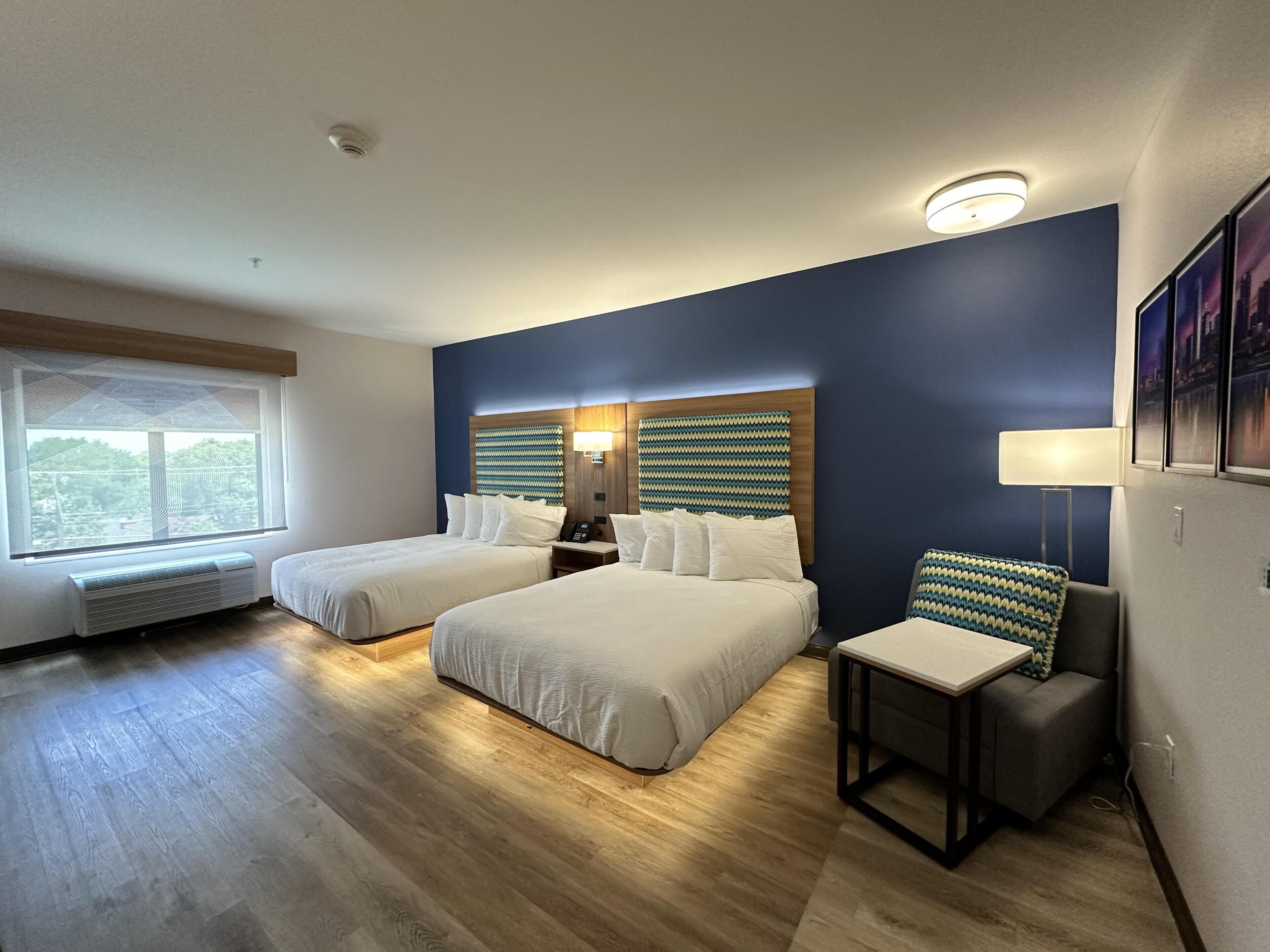 Habitación de hotel con dos camas, un escritorio, una silla, una ventana con persianas, una lámpara de pared y una luz de techo.