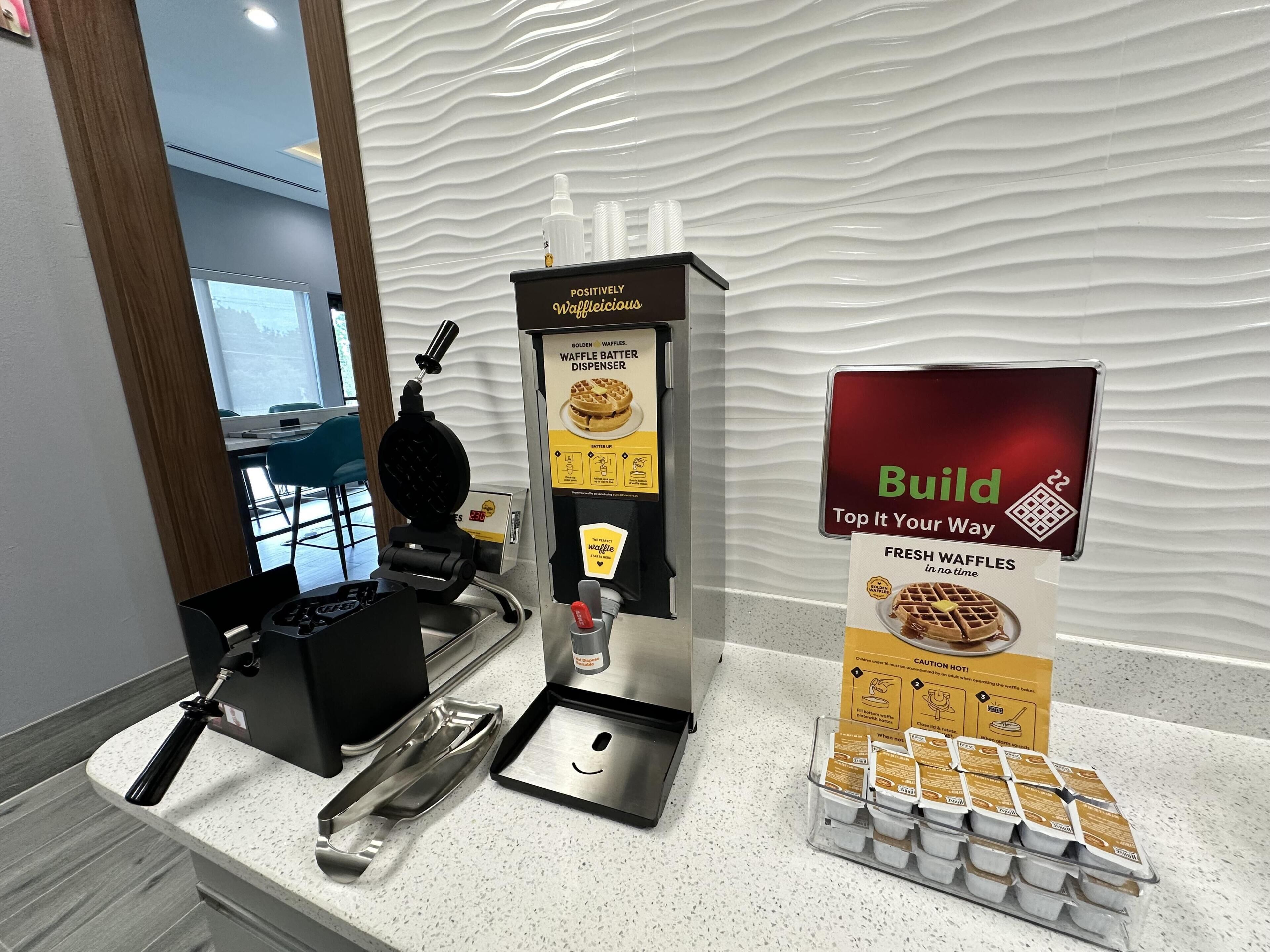 Una máquina para hacer gofres, un dispensador de masa para gofres y un cartel que anuncia gofres recién hechos.