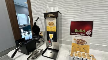 Una máquina para hacer gofres, un dispensador de masa para gofres y un cartel que anuncia gofres recién hechos.