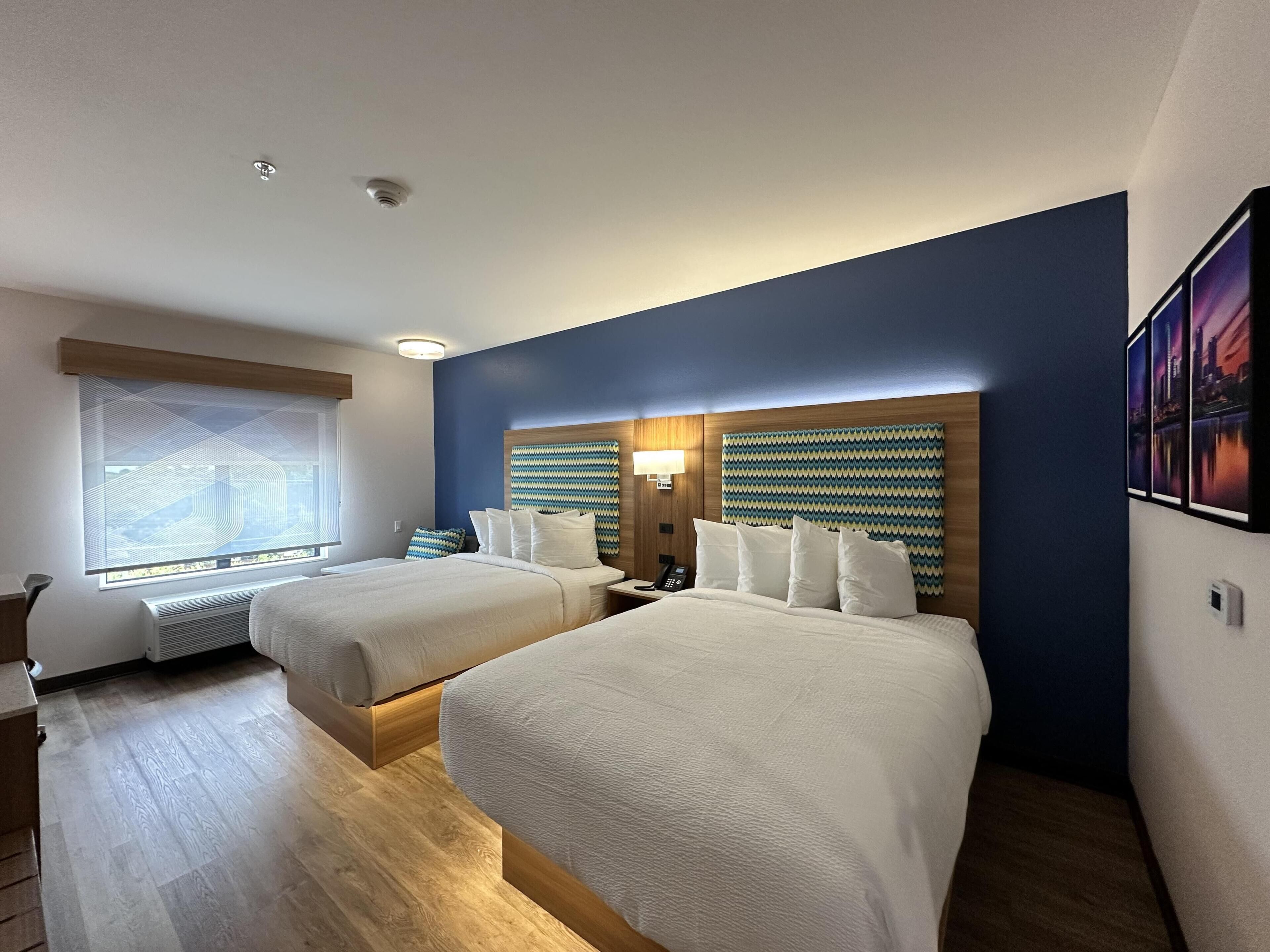 Habitación de hotel con dos camas, una ventana con persianas, una lámpara de pared y una cámara de seguridad en el techo.