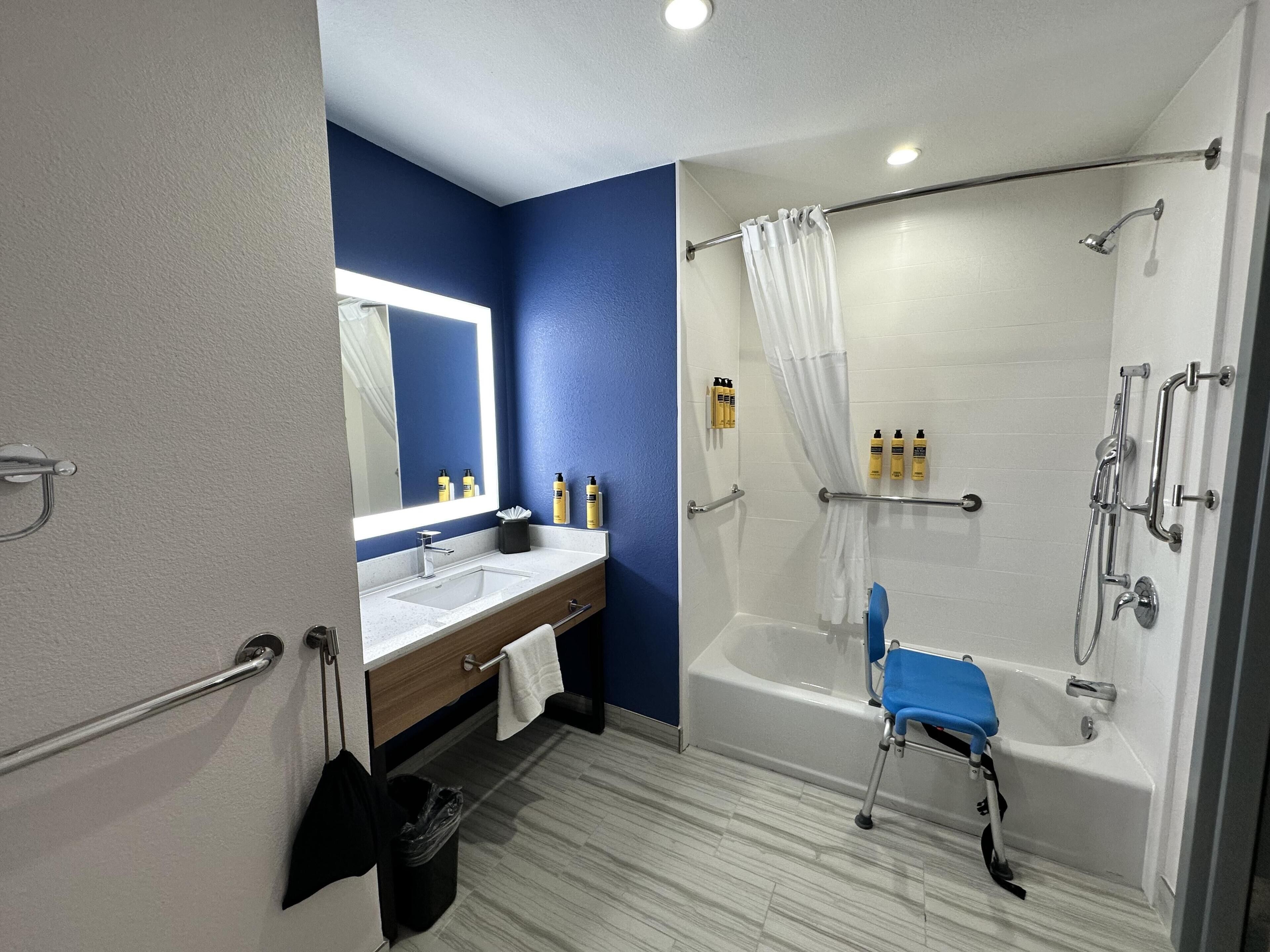Un baño con ducha, lavabo, espejo y silla de ducha accesible para sillas de ruedas.