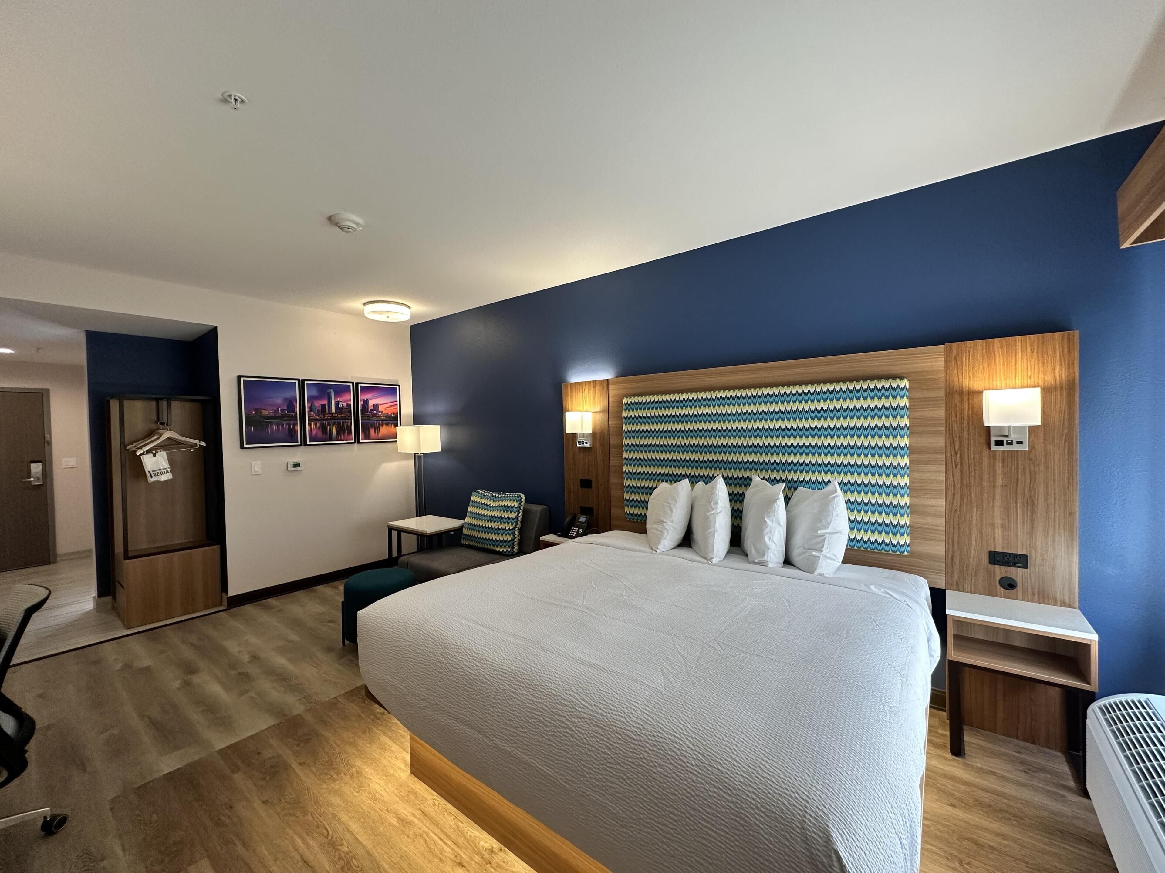 A hotel room with a large bed, a desk, a chair, a desk lamp, a wall-mounted heater, and a door.