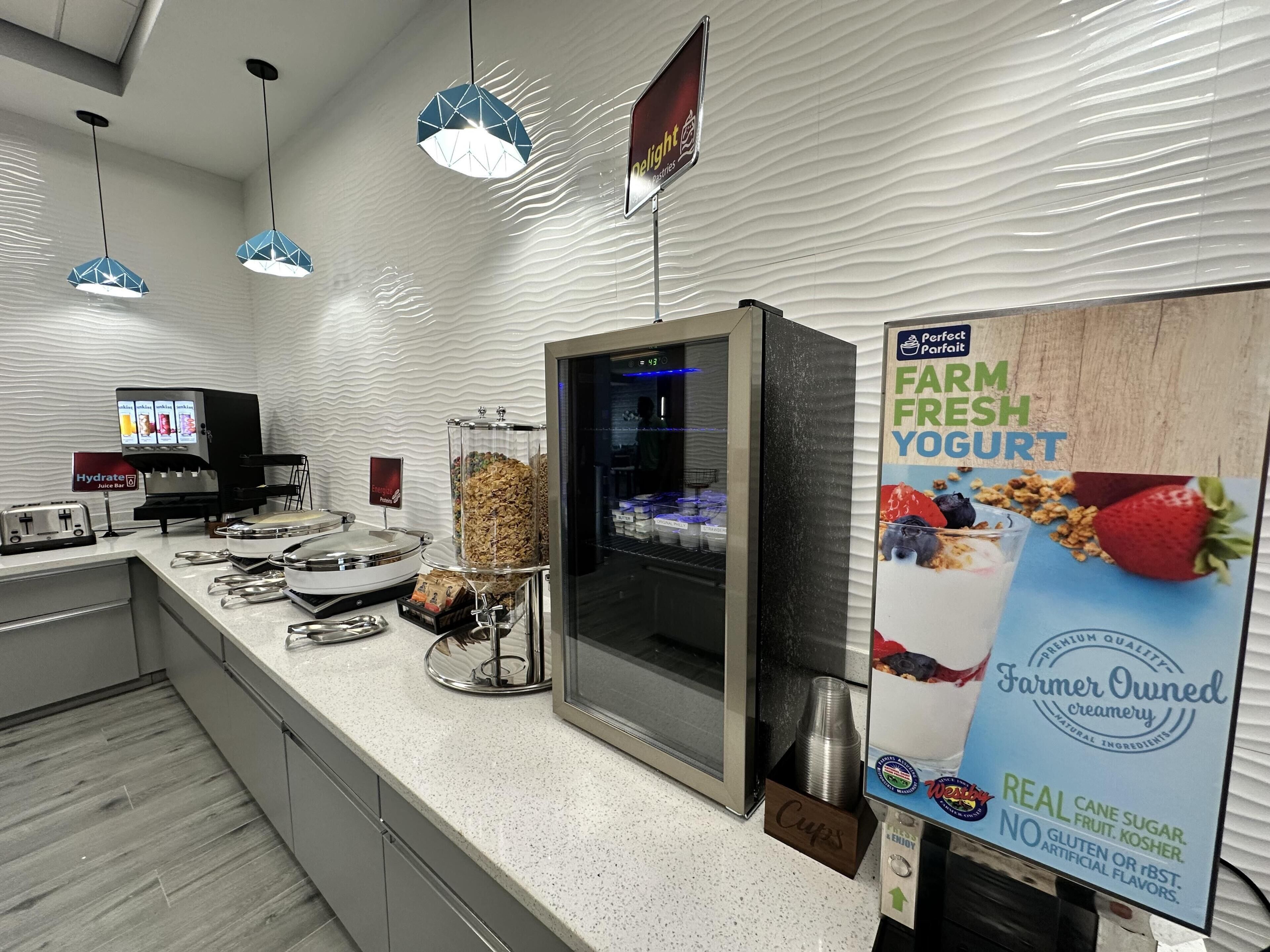 Zona de desayuno autoservicio con mostrador refrigerado, barra de jugos y cartel que anuncia yogur de la granja.