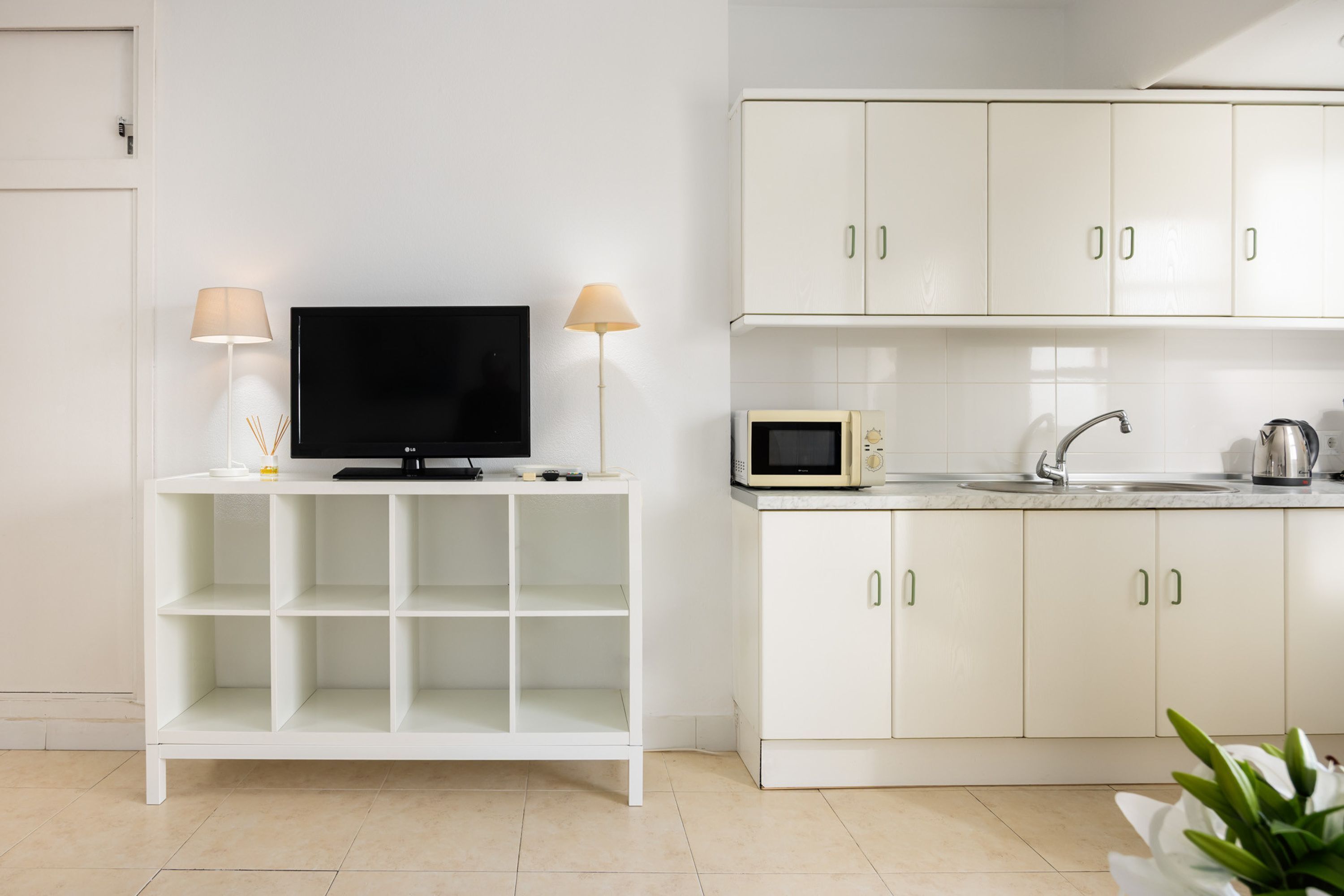Una cocina moderna con armarios blancos, un microondas, un hervidor y un televisor sobre un mueble blanco.