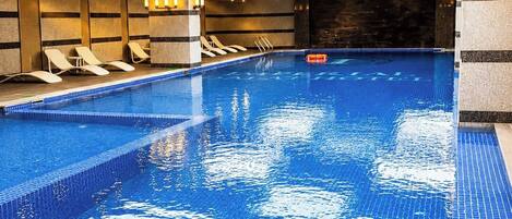 Una piscina interior con agua azul cristalina, vigas de madera en el techo y sillas de descanso a lo largo de la pared.