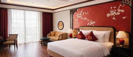 A hotel room with a large bed, a sofa, a chair, a nightstand with a lamp, and a window with curtains.