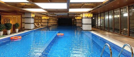 Una amplia piscina interior con agua cristalina y azul, rodeada de sillas de descanso y un techo de madera con tragaluces.