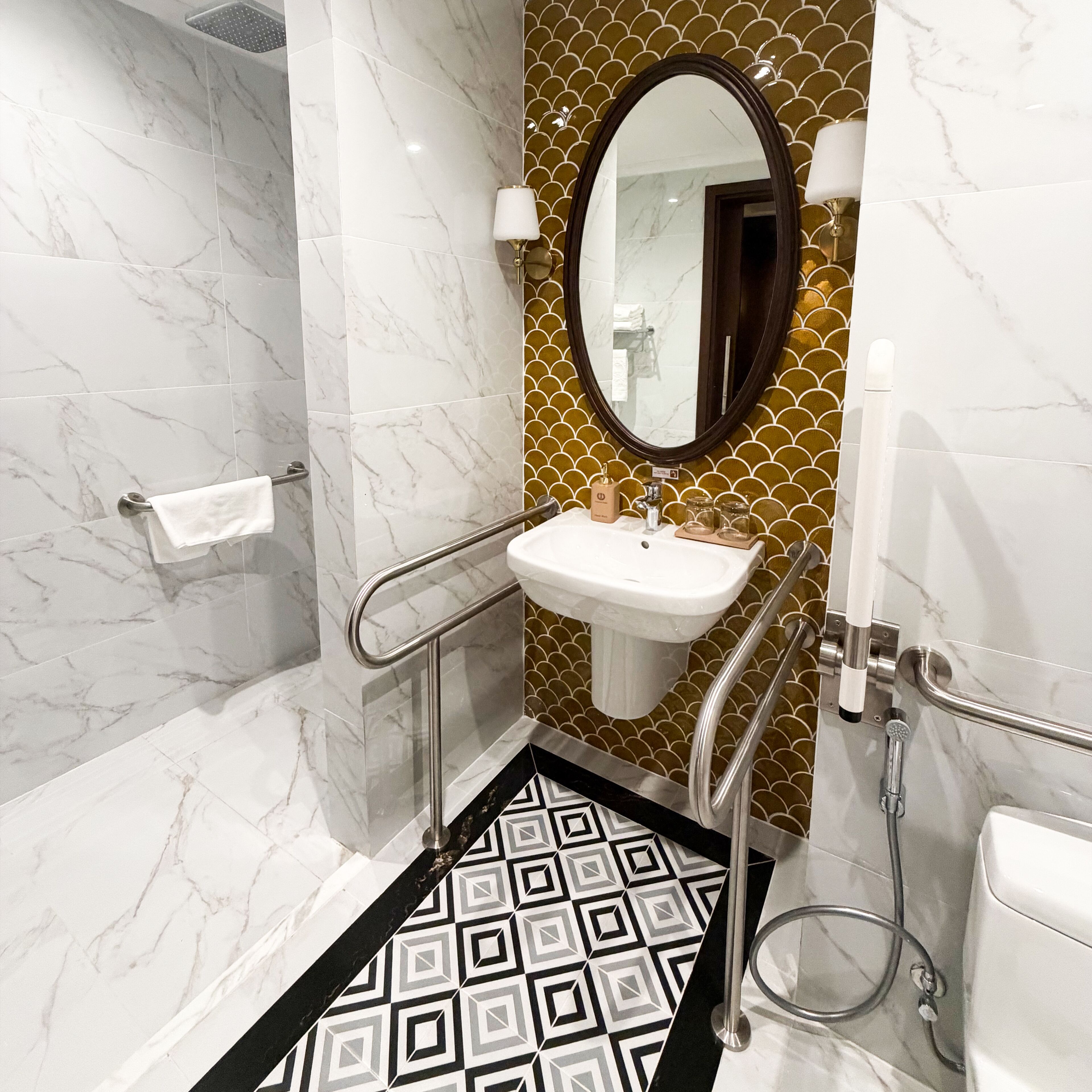 Un baño con piso de mármol, lavamanos con resalte dorado, espejo y zona de ducha.