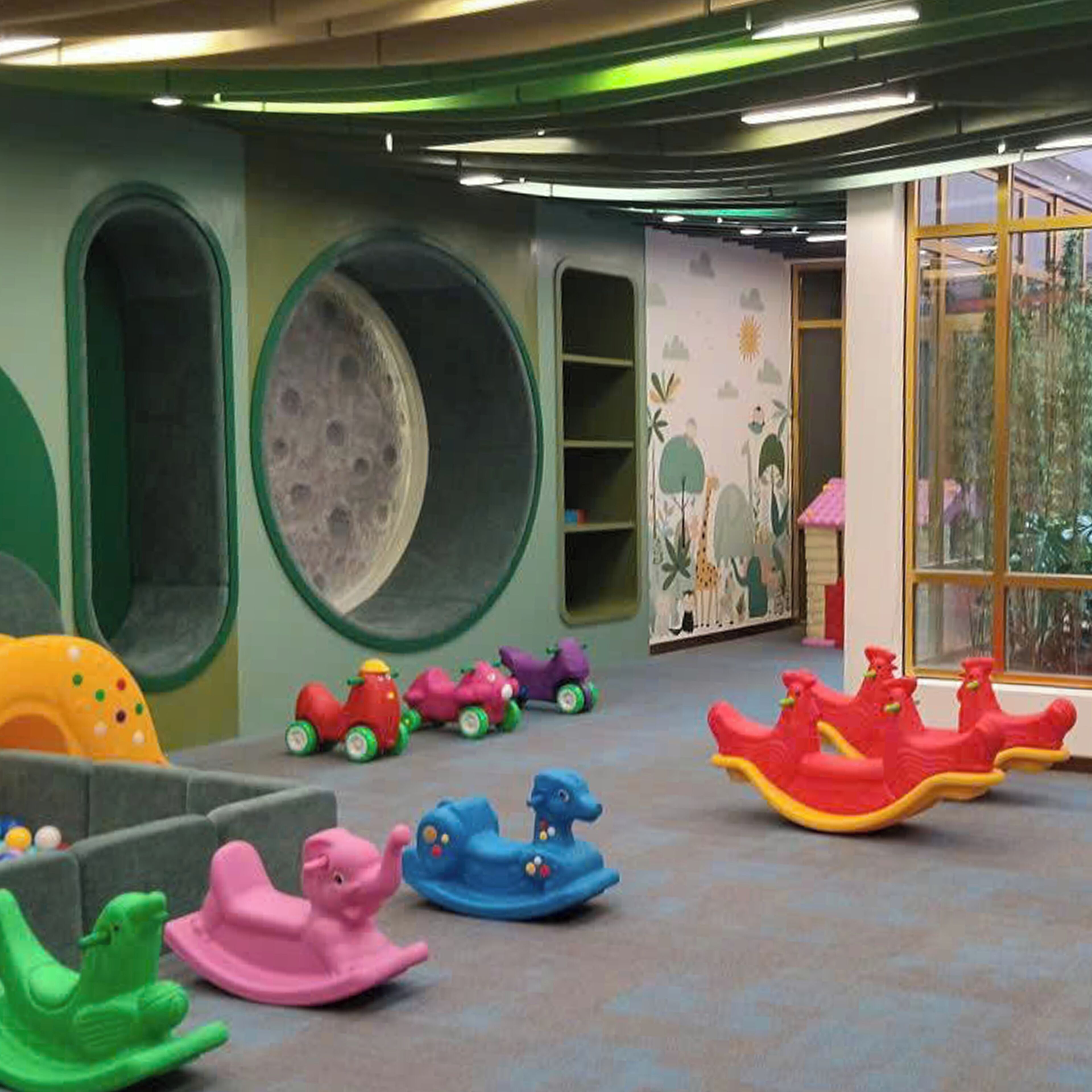 A children's play area with colorful rocking horses, a large moon mural, and a window with a view of trees.