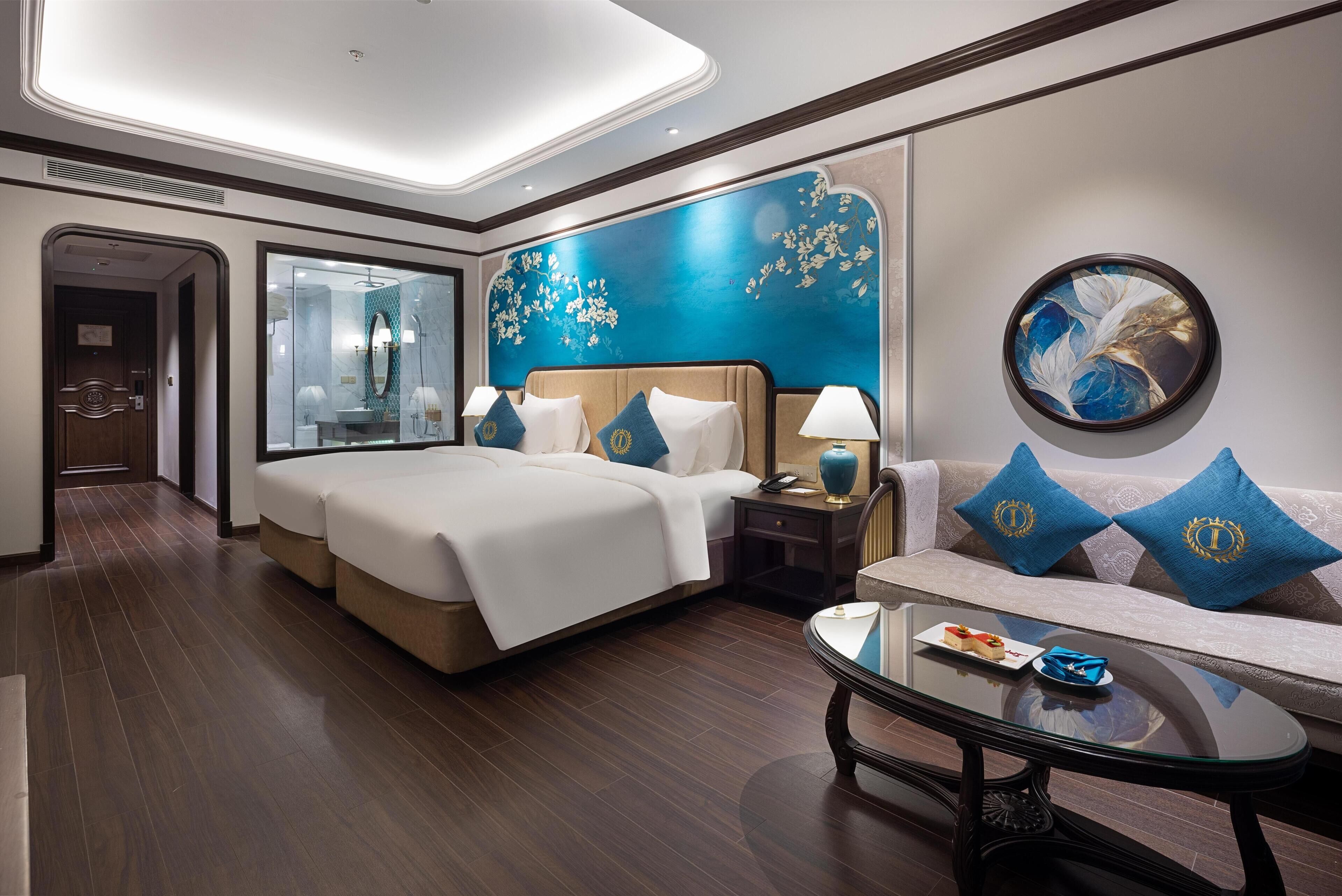Habitación de hotel con una cama grande, un sofá, una mesa de centro de vidrio y una pared decorada con un cuadro floral.