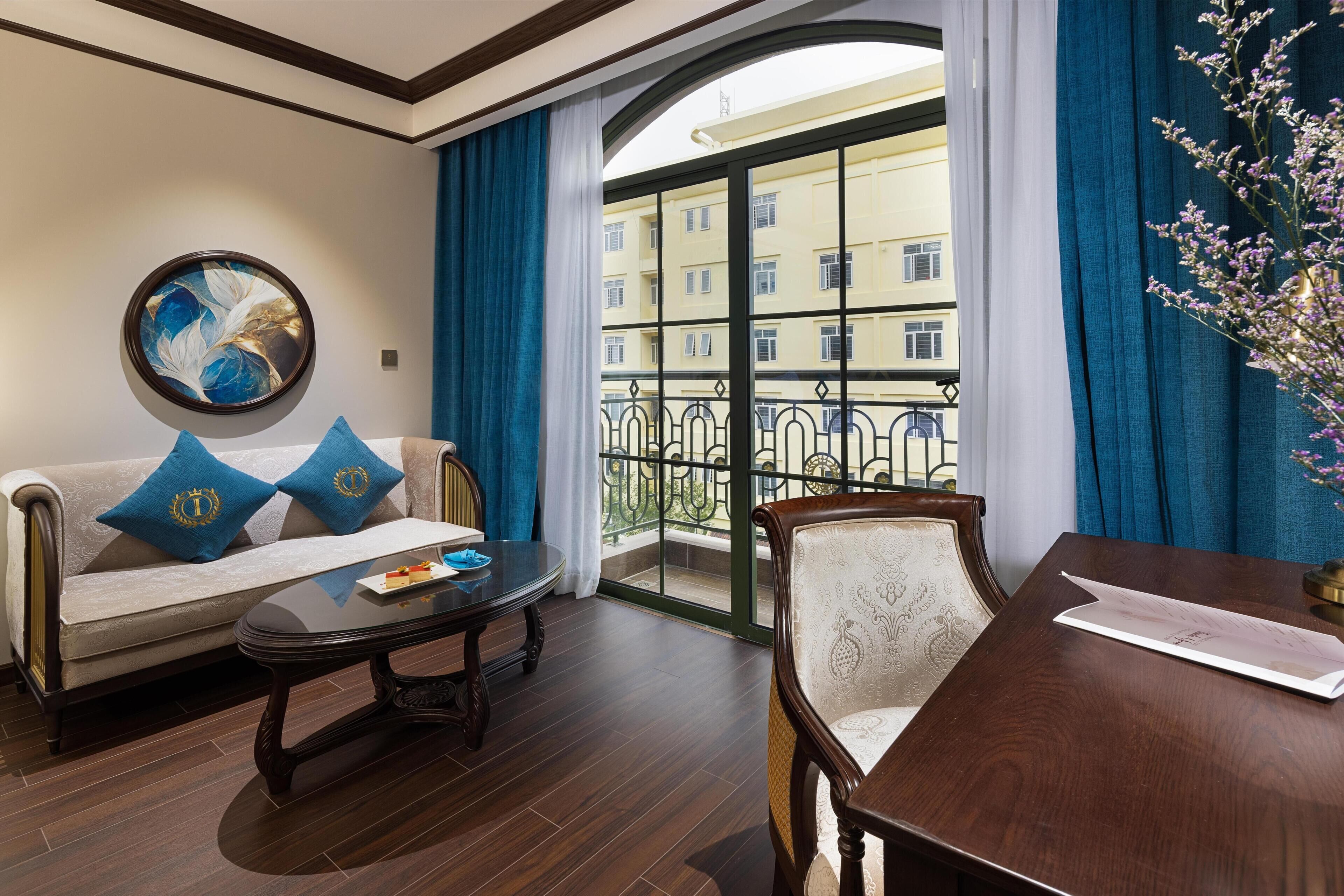A room with a sofa, a table, and a chair. There is a balcony with a railing and a view of buildings.