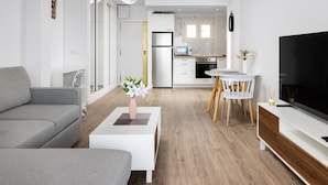 Ein modernes Wohnzimmer mit einem grauen Sofa, einem weißen Couchtisch mit einer Vase, einem Essbereich mit Tisch und Stühlen sowie einer Küche im Hintergrund.