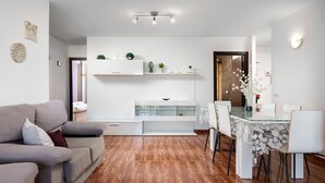 Uma sala de estar moderna com sofá, mesa de jantar com cadeiras, área de cozinha com armário branco e um objeto decorativo na parede.