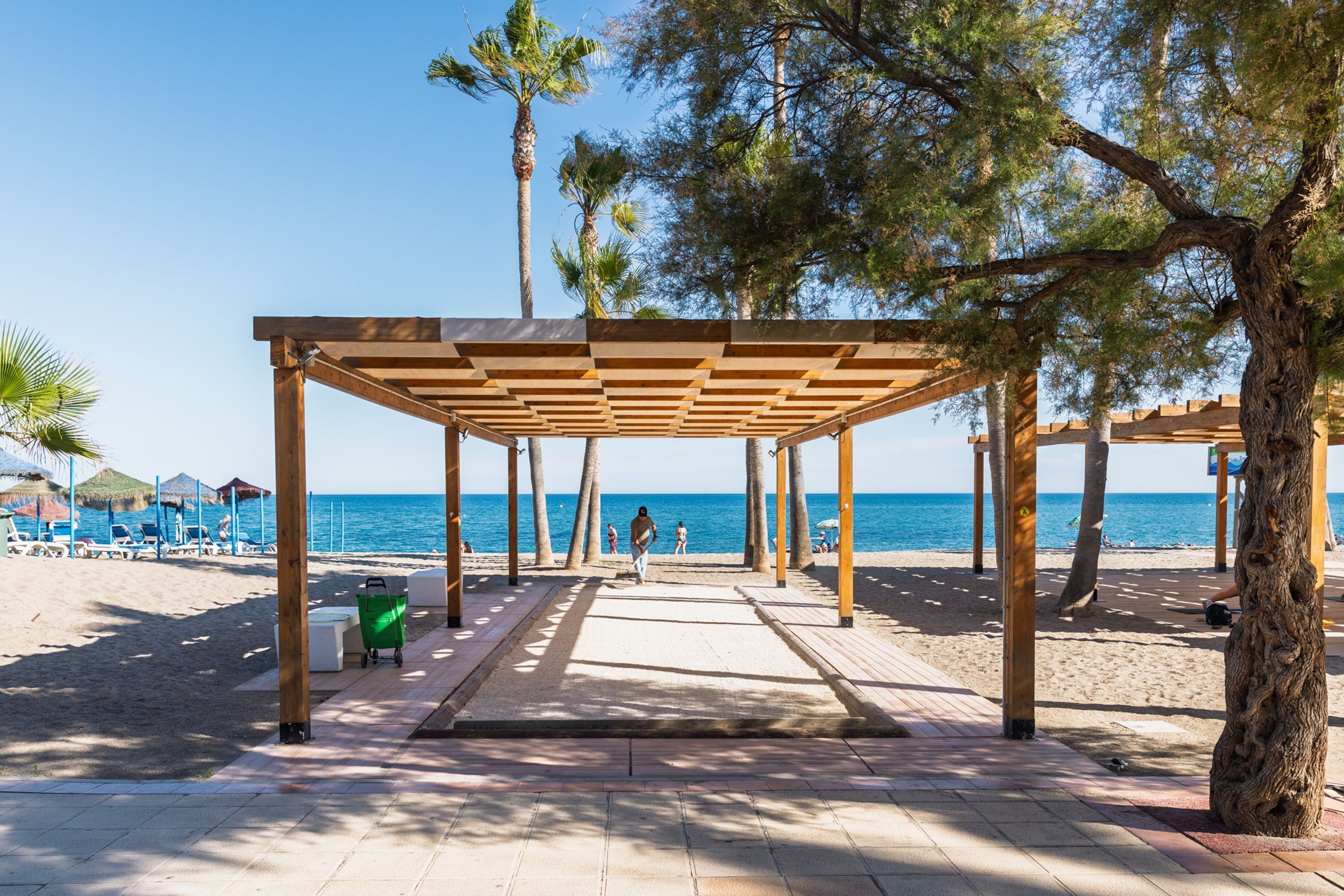 Een promenade aan het strand met houten pergola’s, palmbomen en strandparasols.